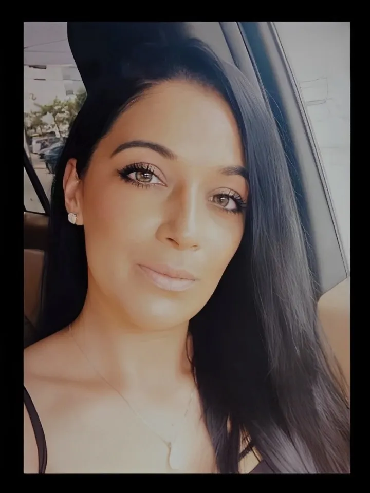Woman with long dark hair and light makeup sitting in a car, looking at the camera.