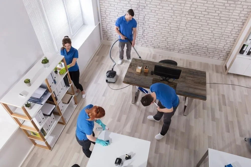 Four people in blue shirts cleaning an office with vacuum, wiping desk, and organizing shelves.